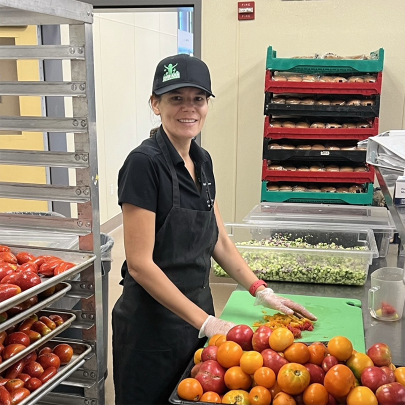 Female food worker cutting up tomatoes with a pile of tomatoes next to her, smiling at the camera.
