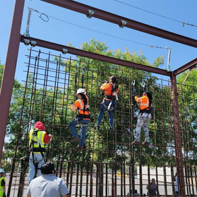 Female construction workers wearing safety harnesses and bright vests climb a rebar wall.