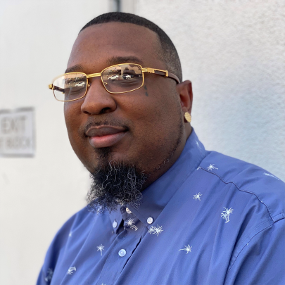 African American man wearing gold glasses and a goatee looking at the camera.