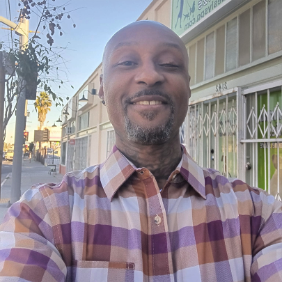 African-American man with goatee takes a selfie and smiles at the camera with a city street behind him.